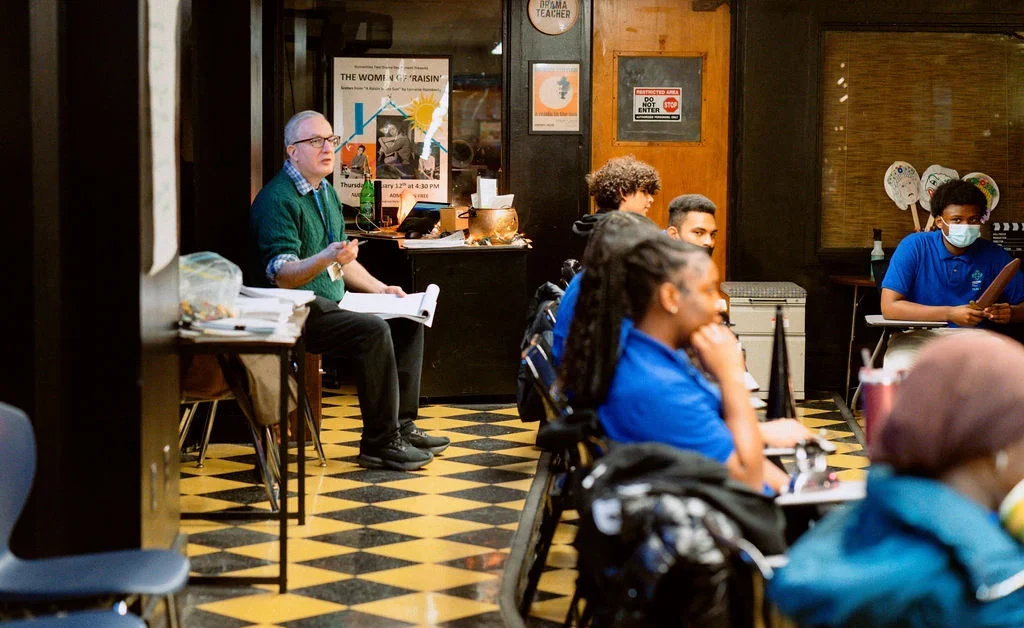 Students and a teacher in a UCHS HUM school sit in chairs and face a speaker who is out of the image.