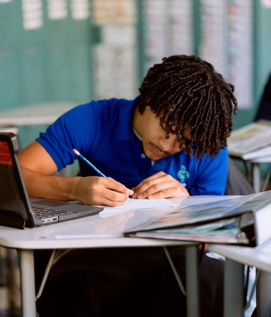 A student at United Charter High Schools in a blue shirt sits at a desk, writing in a notebook with a pencil. An open laptop and textbooks are on the desk.
