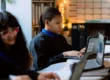 Three United High Schools students sit at desks using laptops in a classroom with a whiteboard and shelves in the background. The focus is on the student in the center.