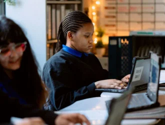 Three United High Schools students sit at desks using laptops in a classroom with a whiteboard and shelves in the background. The focus is on the student in the center.