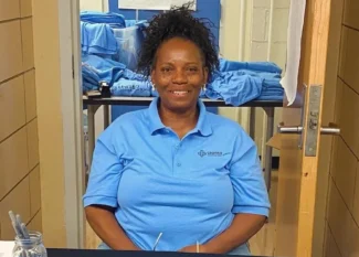 A staff member of United Charter High Schools wears a blue uniform shirt. She is sitting at a table with a selection of blue uniform shirts.