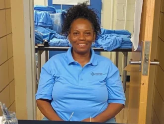 A staff member of United Charter High Schools wears a blue uniform shirt. She is sitting at a table with a selection of blue uniform shirts.