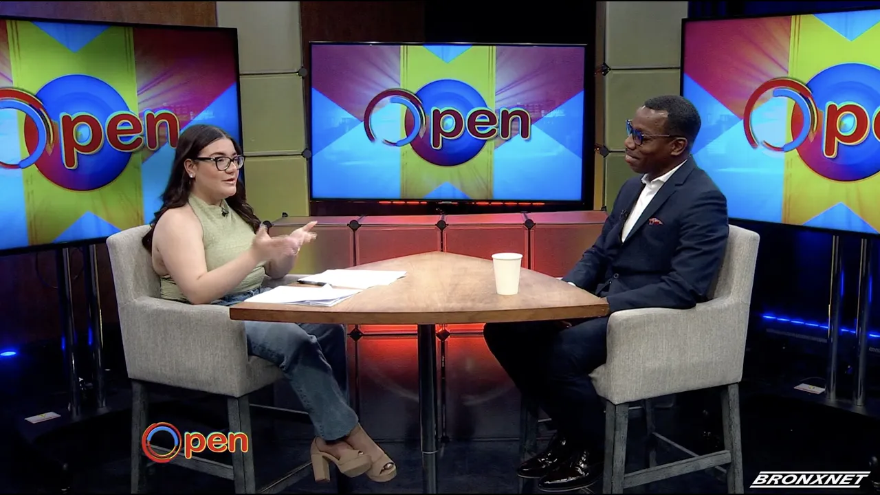 A woman interviews a man in a studio set with "Open" displayed on monitors behind them; both are seated at a table with papers and a cup.