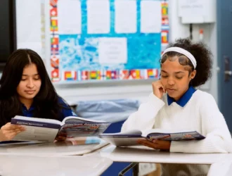 Two students at United Charter High Schools read a textbook while sitting at a table in a classroom.