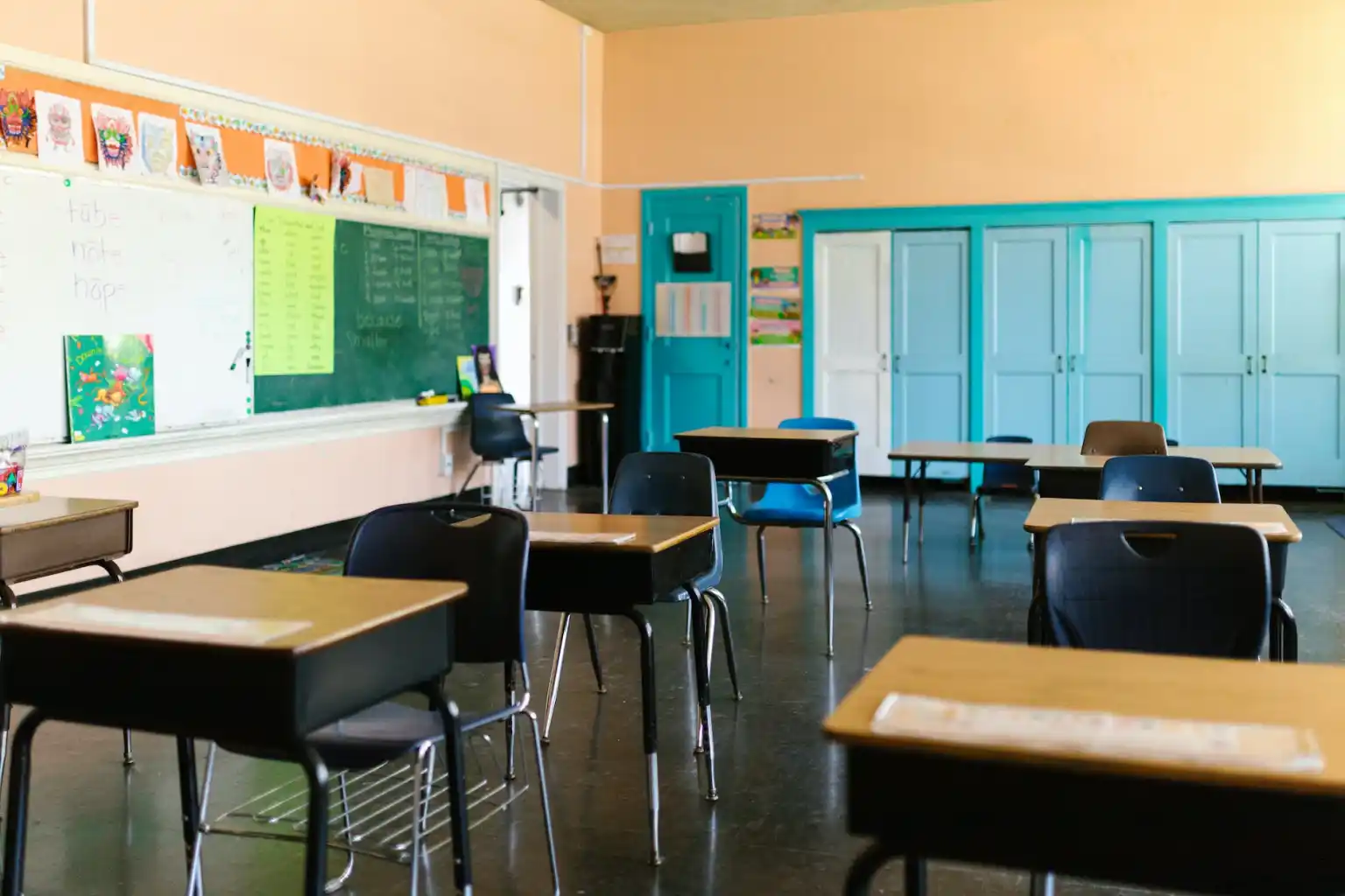 A classroom with empty desks and chairs, a green chalkboard with writing, bright walls, and blue cabinets along the back wall.