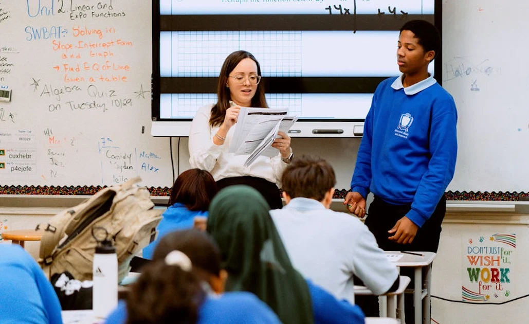 A teacher stands at the front of a classroom holding papers, speaking to students, as another student stands beside her; classmates face forward at their desks, embodying the spirit of rigorous education NYC is known for.