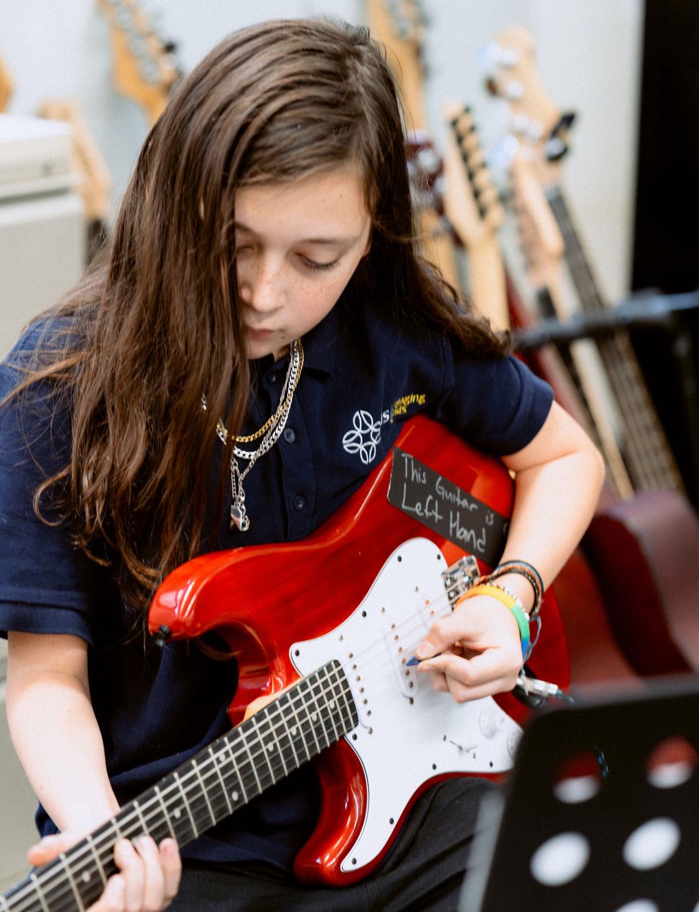 A young person with long hair plays a red electric guitar labeled "This Guitar is Left Handed" in a room with several guitars in the background.