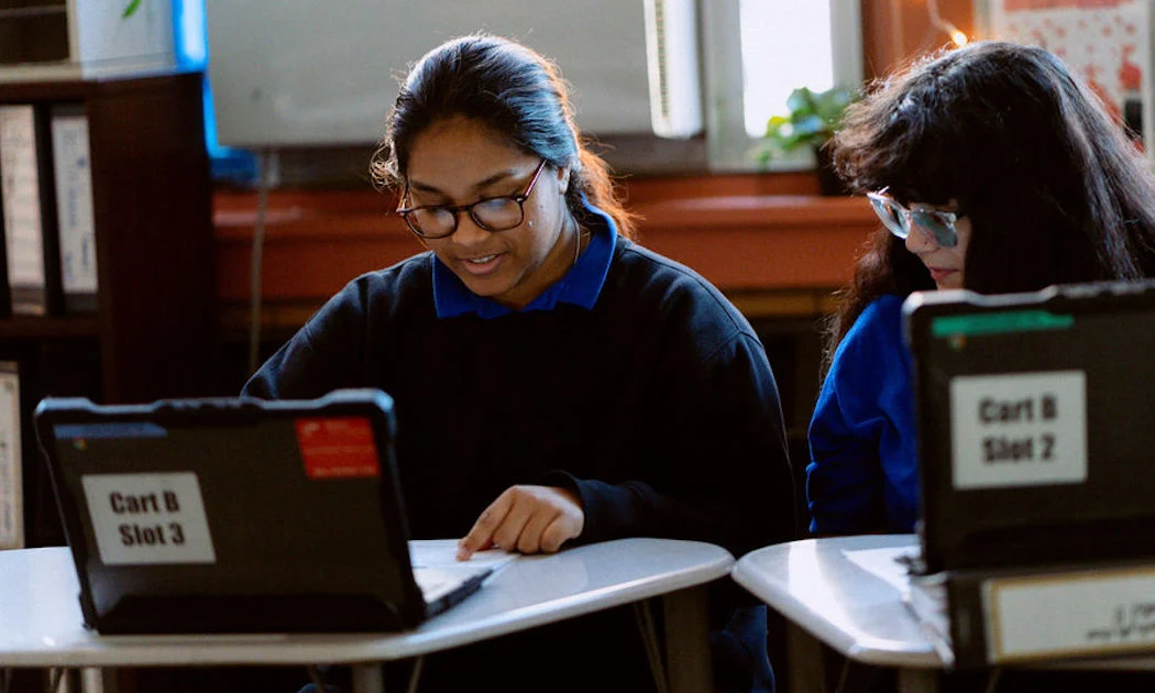 Two students in a classroom benefit from a rigorous education NYC is known for, using laptops labeled "Cart B Slot 3" and "Cart B Slot 2" as one points to a paper on the desk.