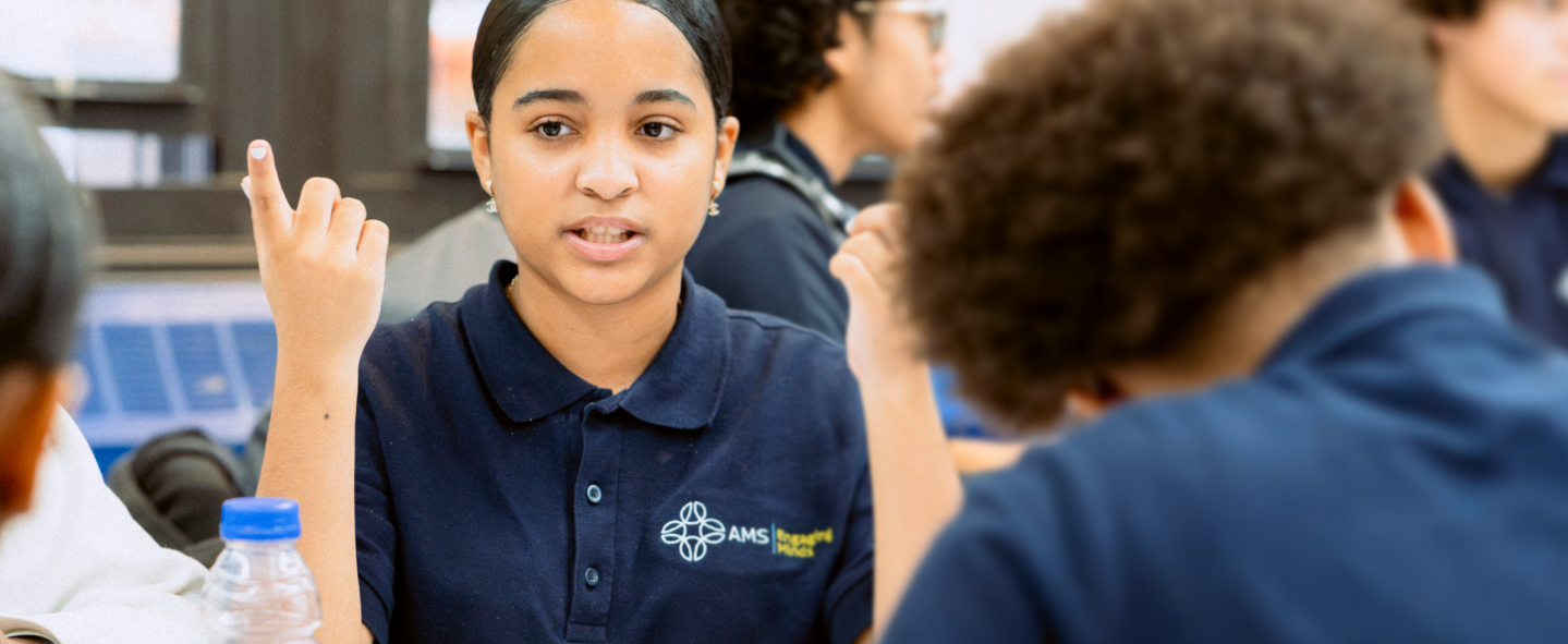 A student in a navy blue school uniform raises her hand while talking in a classroom, with other students visible in the background.