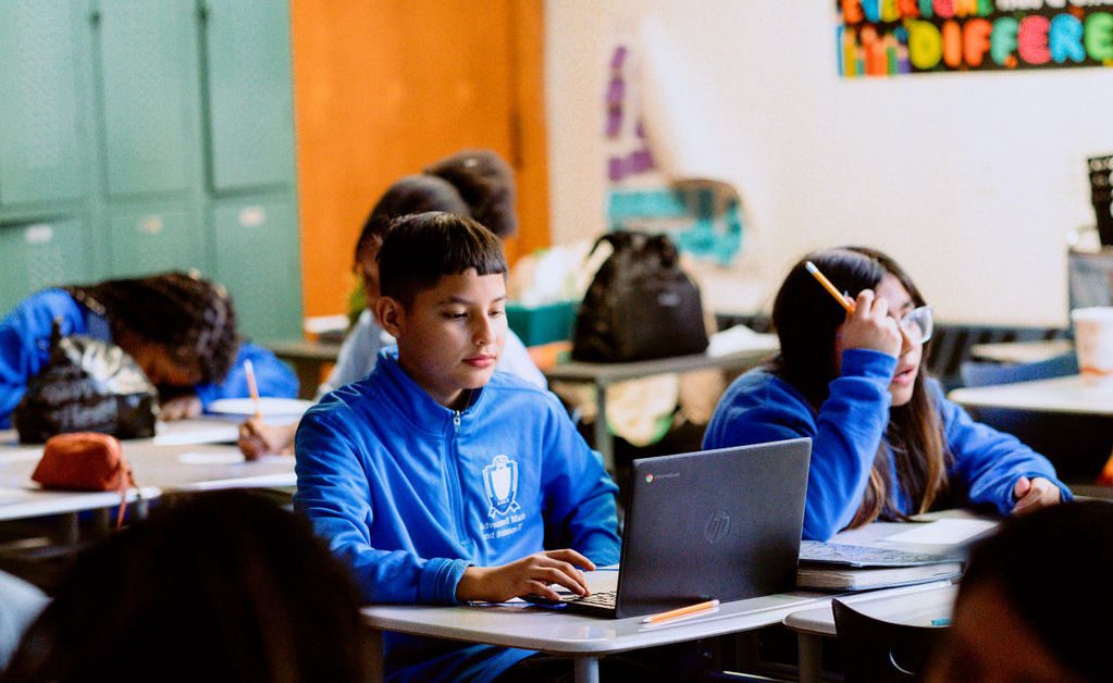 Students in blue uniforms sit at desks in a classroom; one is using a laptop while others write on paper.