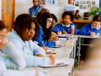 Students in blue uniforms sit at desks in a classroom, writing in notebooks, with one student in the center looking toward the camera.