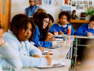 United Charter Students in blue uniforms sit at desks in a classroom, writing in notebooks, with one student in the center looking toward the camera.