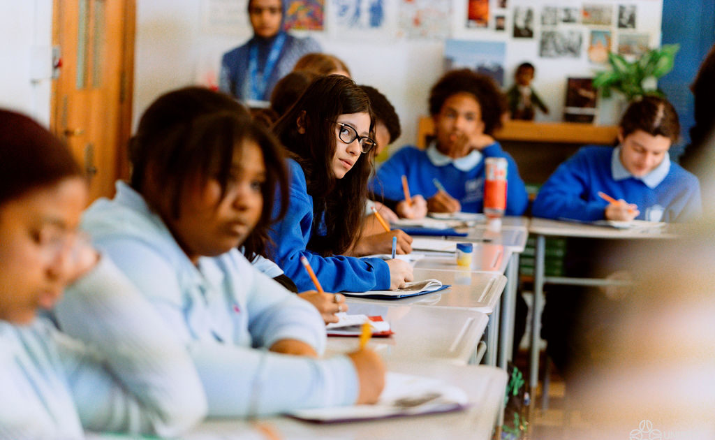 United Charter Students in blue uniforms sit at desks in a classroom, writing in notebooks, with one student in the center looking toward the camera.