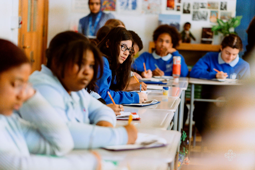 Students in blue uniforms sit at desks in a classroom, writing in notebooks, with one student in the center looking toward the camera.