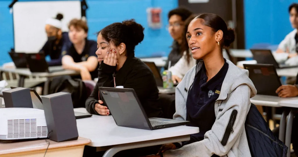 Students sit at desks in a classroom, using laptops and listening attentively. One student in the foreground is speaking, while others focus on their screens or pay attention to the lesson.
