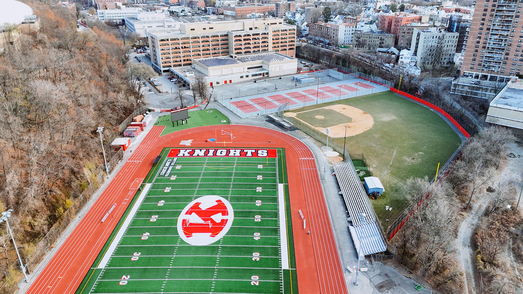 Aerial view of a sports complex featuring a football field marked "KNIGHTS," a running track, tennis courts, a baseball field, bleachers, and surrounding buildings.