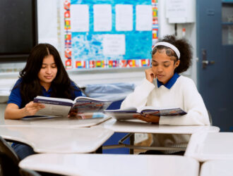 Two students sit at desks in a classroom, reading textbooks. The classroom has posters, a bulletin board, and a closed door in the background.
