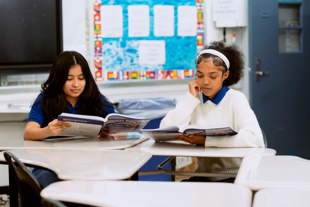Two students sit at desks in a classroom, reading textbooks. The classroom has posters, a bulletin board, and a closed door in the background.