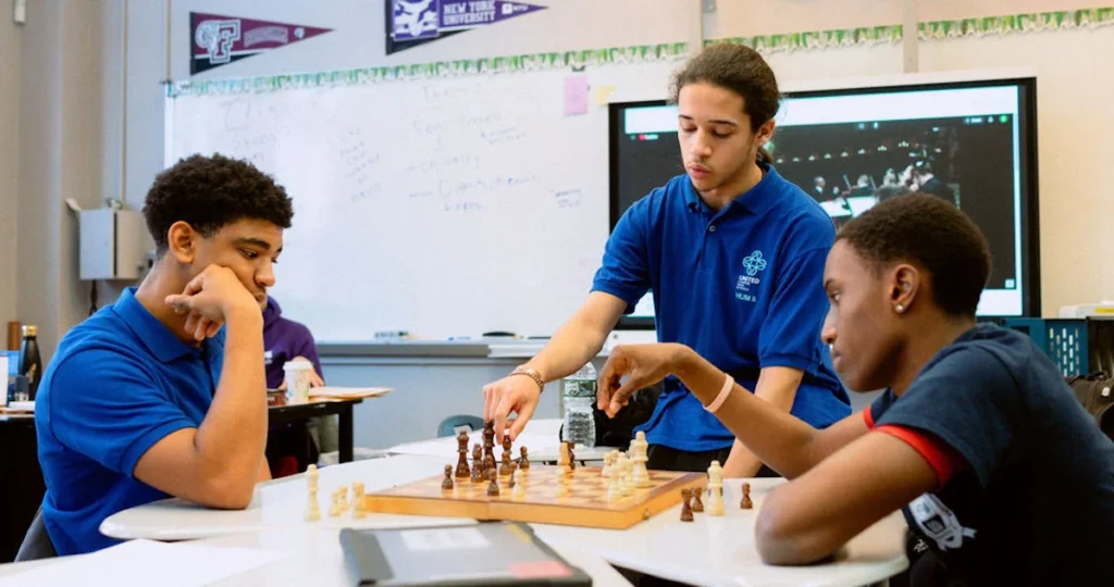Three students in blue shirts play chess in a classroom; one is making a move while the others watch and think. A whiteboard and a screen are visible in the background.