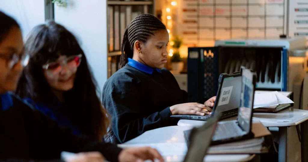 Three students sit at desks in a classroom, working on laptops and surrounded by folders and papers.