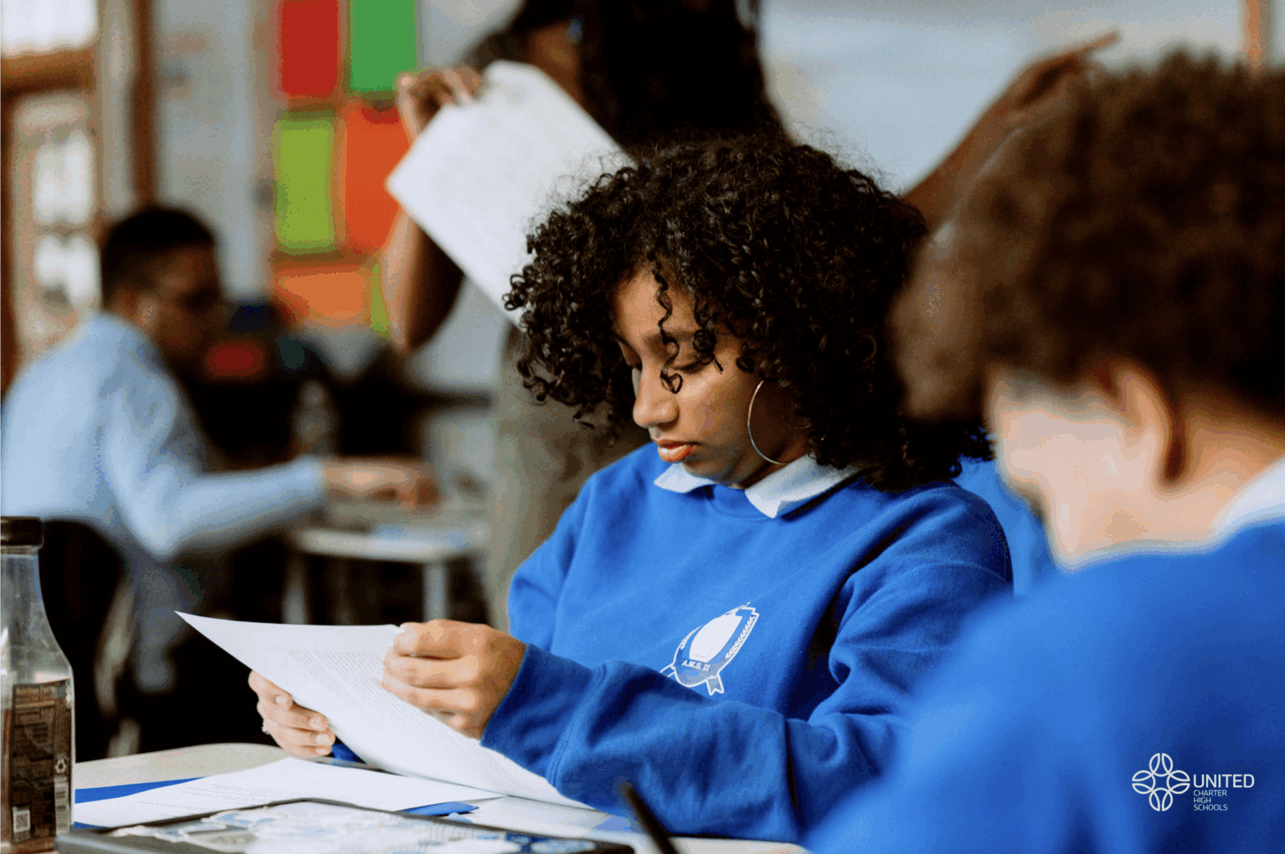 A student in a blue school uniform reads a sheet of paper at her desk in a classroom setting.