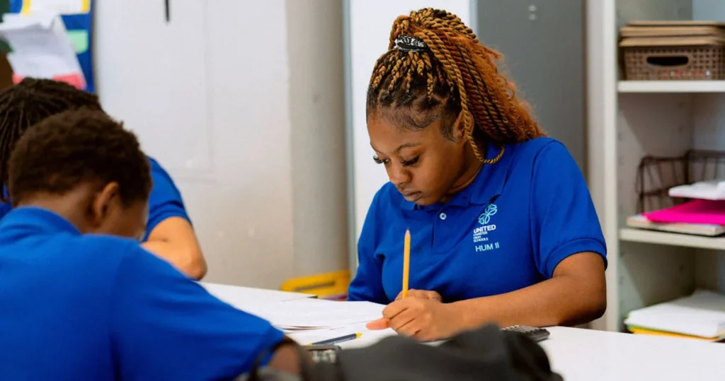 A student in a blue polo shirt writes on paper at a desk in a classroom, with other students seated nearby.