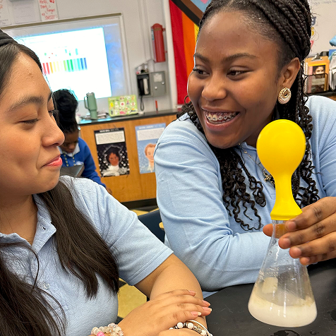 Two students in a classroom smile while one holds a flask with a yellow balloon on top, likely participating in a science experiment.