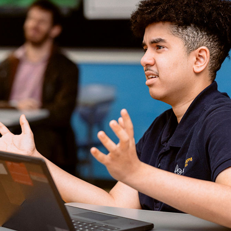 A student with curly hair sits at a desk with a laptop, gesturing with open hands and an expressive face, while another person is seated in the blurred background.