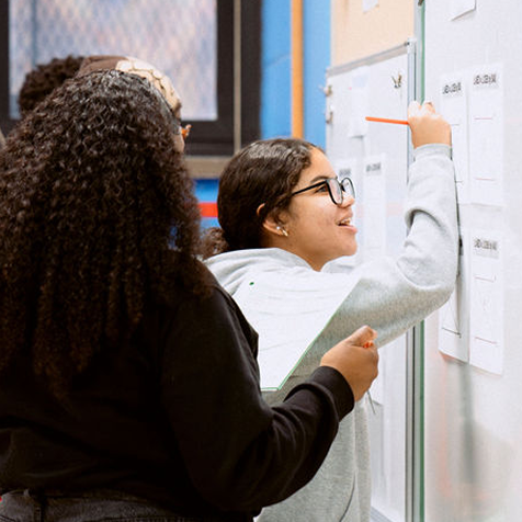 Two students stand at a whiteboard, with one writing on it using a red marker while the other observes closely.
