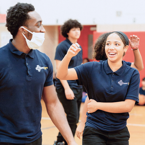 A group of young people in navy blue polos interacts in a gymnasium; one person wears a mask, and another smiles with an arm raised.