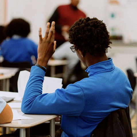 A student wearing a blue jacket sits at a desk in a classroom, raising their hand, with an instructor standing in the background.