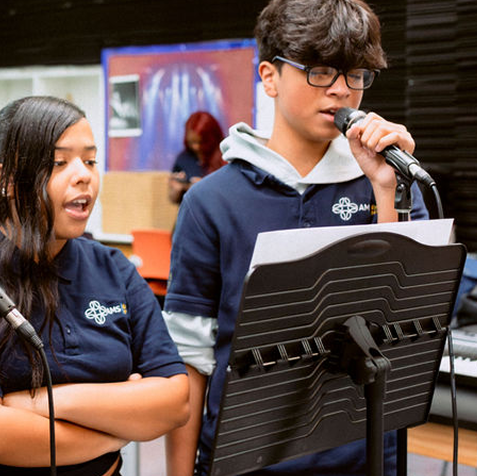 Two students in matching navy shirts stand at microphones, singing from sheet music on a stand in a classroom or music room setting.