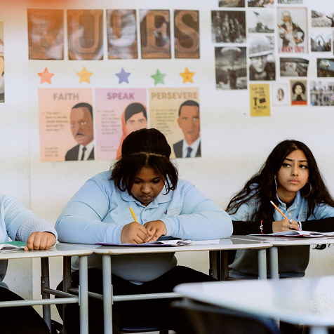 Three students sit at desks in a classroom, writing in notebooks. Educational posters and photos are displayed on the wall behind them.