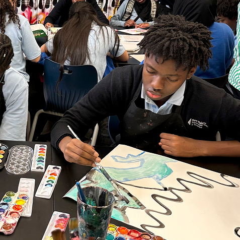 A student sits at a table painting a large abstract design with watercolors, surrounded by paint supplies and other students working on similar art projects.