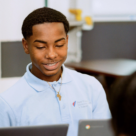 A young man in a light blue polo shirt sits indoors and looks down at a laptop, smiling slightly.