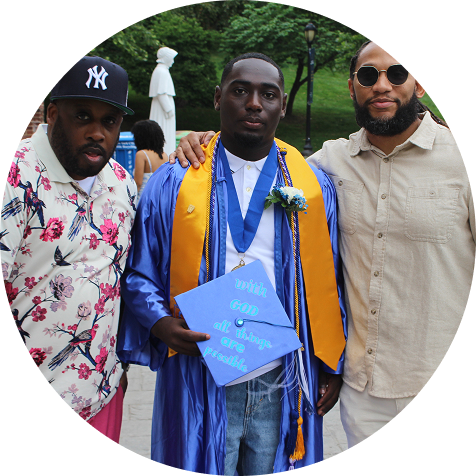 Three men pose together outdoors; the center one wears a graduation cap and gown, holding a decorated cap that reads "with God all things are possible.