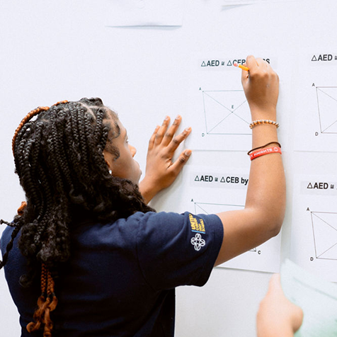 A student with braided hair writes on a whiteboard covered with printed geometry diagrams and labeled triangles.