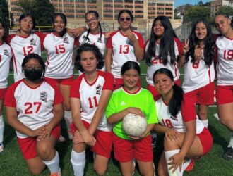 A girls’ soccer team poses together on a field in red and white uniforms, with one player holding a soccer ball at the front.
