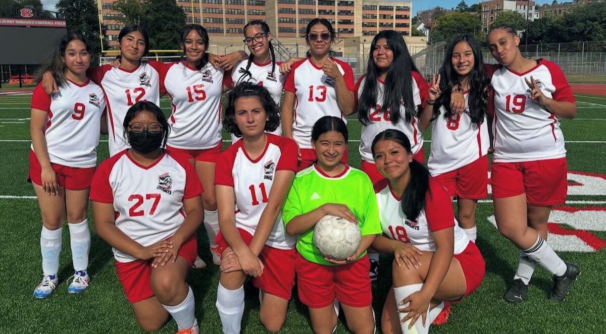 A girls’ soccer team poses together on a field in red and white uniforms, with one player holding a soccer ball at the front.