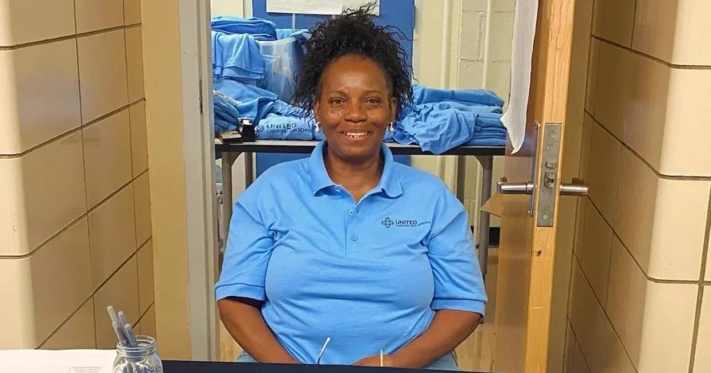 A woman wearing a light blue polo shirt sits at a table in a hallway with folded shirts stacked on a cart behind her.