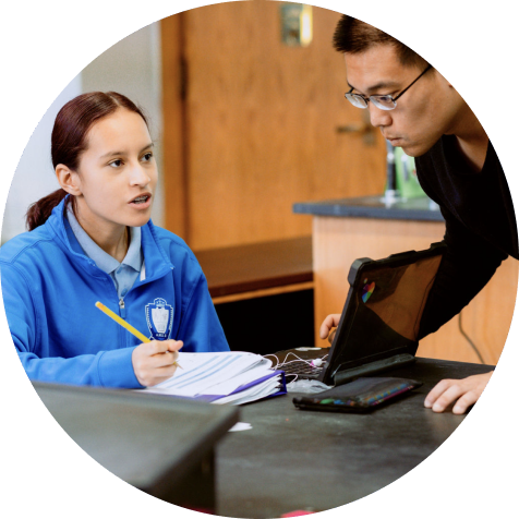 A student in a blue jacket talks to a teacher while seated at a desk with a notebook, pencil, and tablet.
