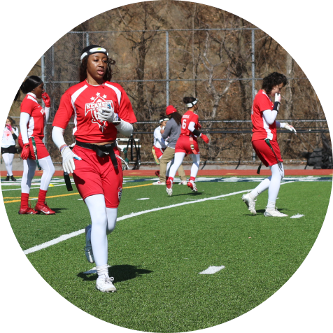 A group of athletes in red uniforms play flag football on a green field, with some running and others preparing for the next play.