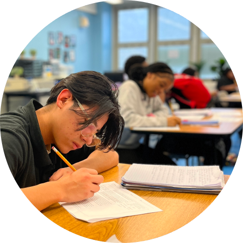 Student sitting at a desk writes on a sheet of paper with a pencil in a classroom; other students are also working at their desks in the background.
