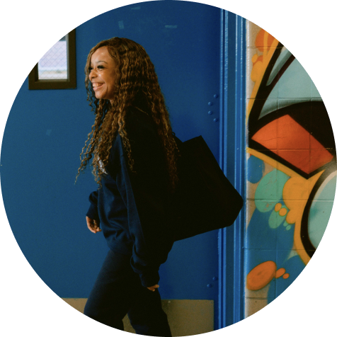 A woman with long curly hair, wearing dark clothing and carrying a black bag, walks past a blue wall with colorful graffiti art.