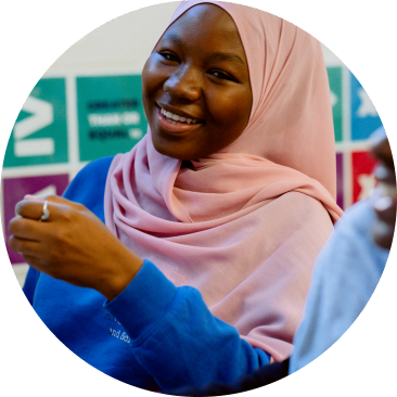 A person wearing a pink hijab and blue shirt smiles while sitting indoors, with colorful charts visible on the wall behind them.