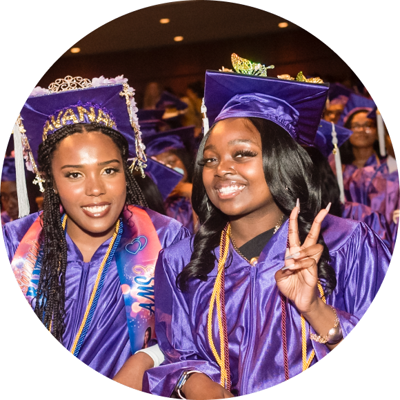 Two graduates in purple caps and gowns smile at the camera; one holds up a peace sign during a graduation ceremony.