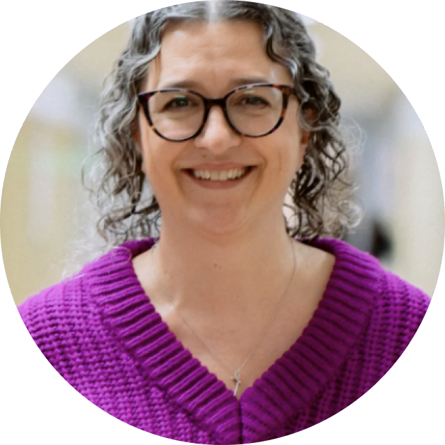 A person with curly gray hair, glasses, and a purple sweater smiles while standing indoors.