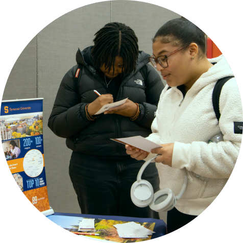 Two students stand at a table with papers and brochures. One is writing on a notepad; the other holds papers and headphones. A Syracuse University poster is displayed in the background.