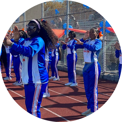 A group of cheerleaders in blue and white uniforms perform a routine on a track near a chain-link fence decorated with blue balloons.