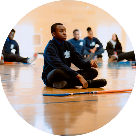 A group of people sit on a gym floor in athletic wear, with hockey sticks placed in front of them. The focus is on one person sitting cross-legged in the foreground.